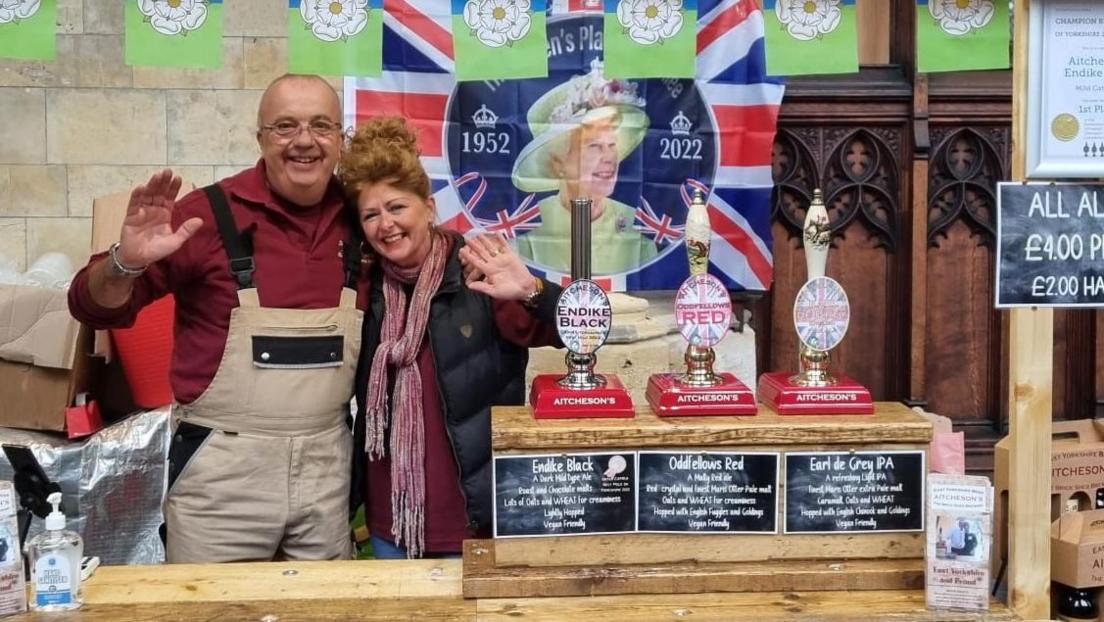 A man and a woman wave and smile as they stand behind a stall at a beer festival in front of a Union flag with a picture of Queen Elizabeth II. He wears glasses, a red shirt and an apron stands. She wears a black gillet and red scarf. Their still includes beer pumps and labels.