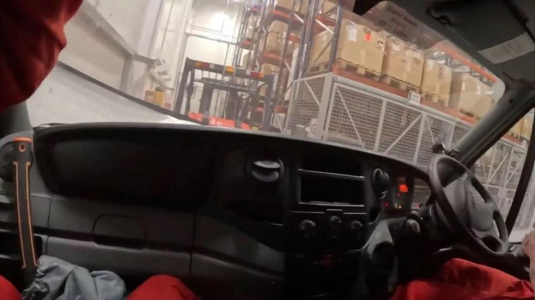 Two people wearing full red boiler suits sitting inside a forklift and riding around the warehouse at the Elbit Systems HQ. There are tall metal shelves stacked full of cardboard boxes on wooden crates. 