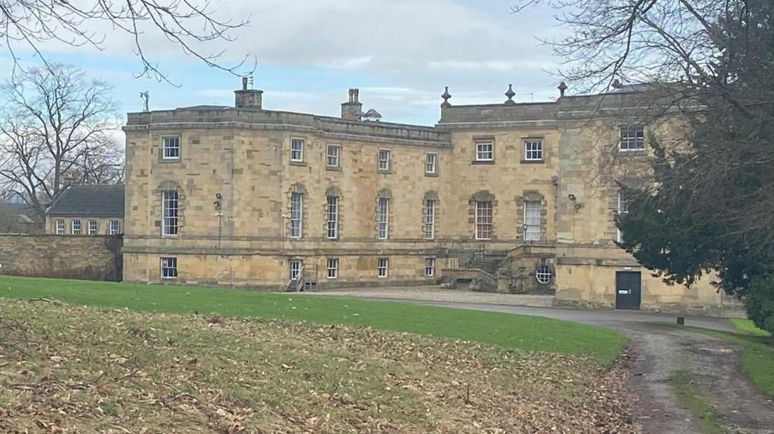 An exterior shot of Gilling Castle in North Yorkshire, showing the front and a lawn covered in leaves. The grand building has several tall windows and a grand flight of stairs. 