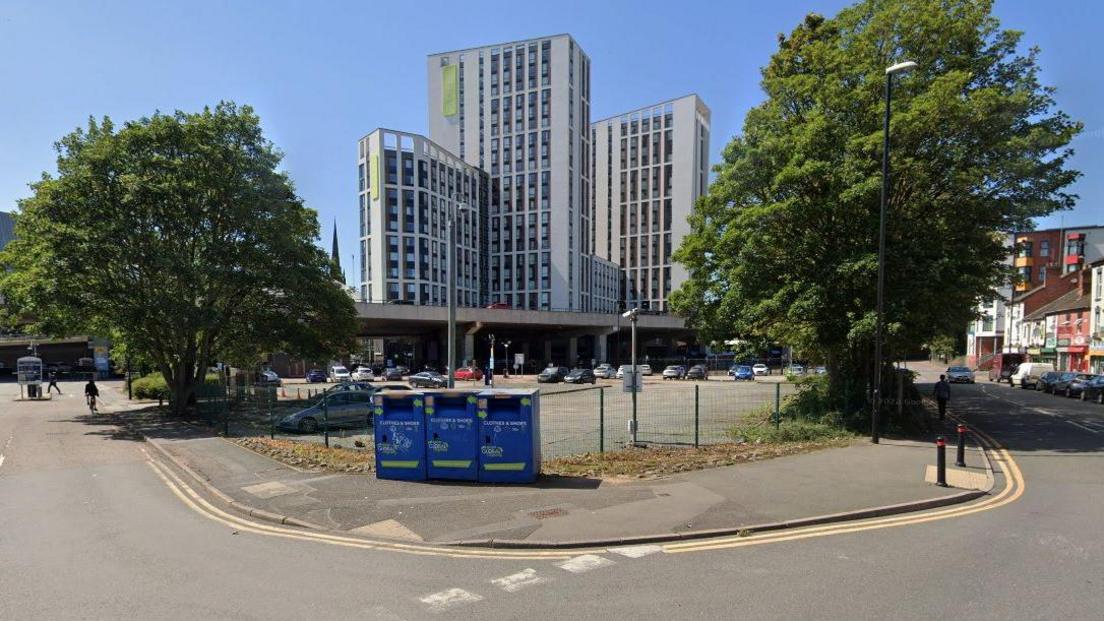 A Google street view image of a car park behind a fence on the corner of two roads. There are two trees either side of the car park and white towers in the background. A ring road is visible just behind and above the car park.