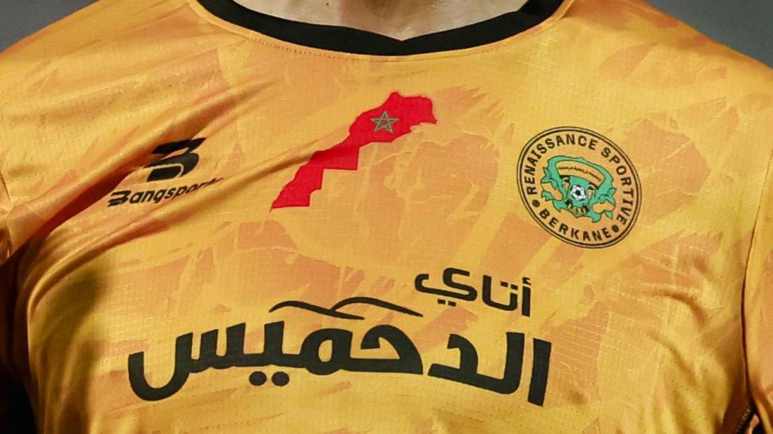 Detail of a RS Berkane football shirt, showing an extended map of Morocco in red on the chest of an orange shirt, in between a sponsor's logo and the club badge