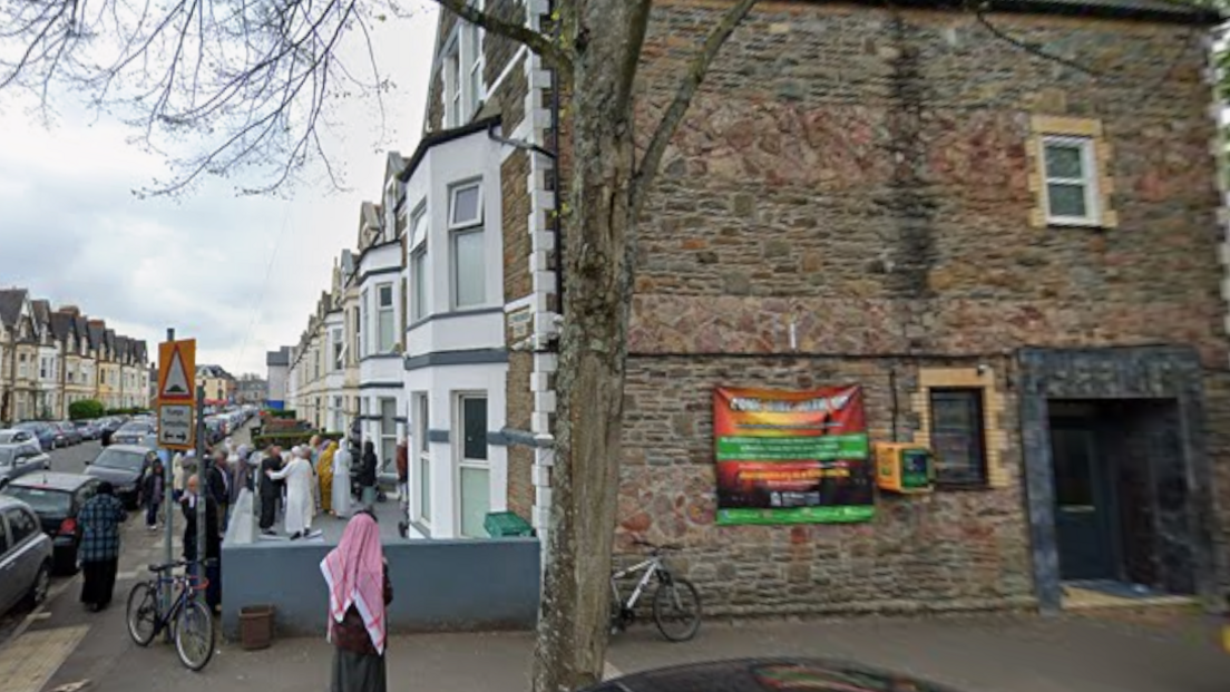 A google street view image of the corner where the mosque is. It is a reddish brown brick building at the end of a row of homes all with white framed windows in the front facing the streets. There is a person in a red abaya standing in front of the building