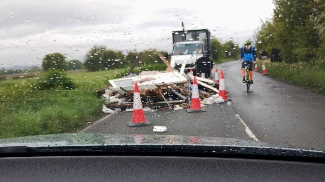 A large pile of what appears to be building waste is dumped across a narrow, rural road. It has been cordoned off, as one cyclists rides past it.