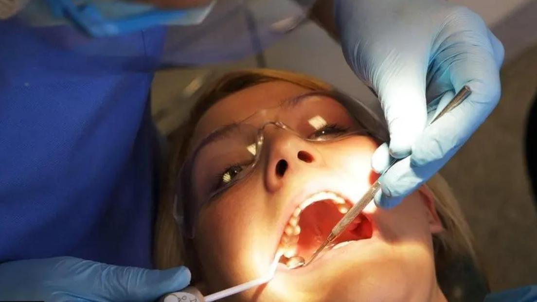 A close up of a patient's face sitting in dentist's chair wearing goggles with a dentist tool in their mouth