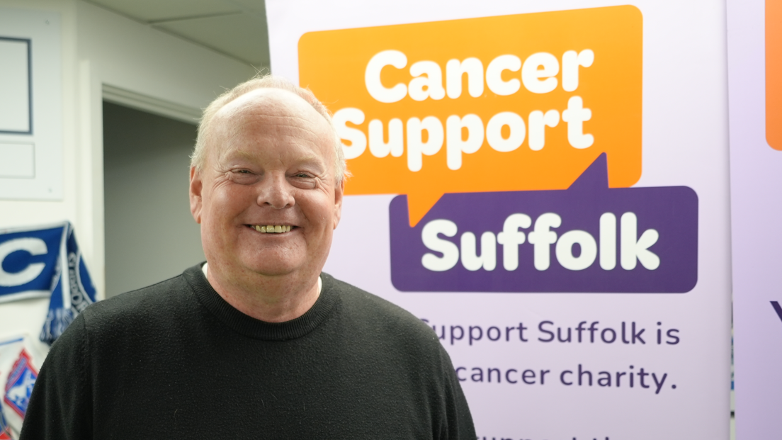 A head and shoulders image of Mark Murphy. He is smiling and standing in front of a back drop featuring the Cancer Support Suffolk logo. He is wearing a green jumper and looking into the camera.