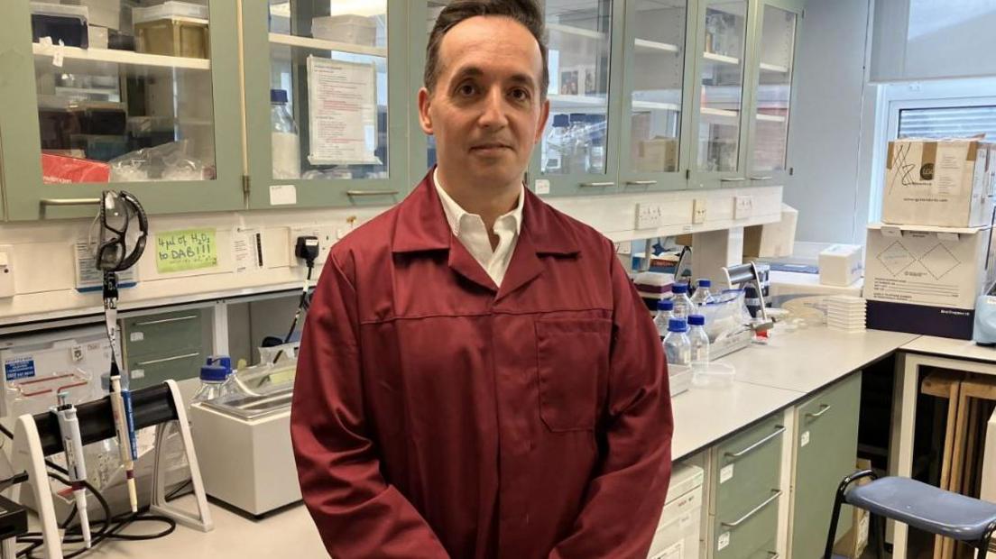 Dr Nik Georgopoulos from Sheffield Hallam University. A dark haired man in a dark red lab coat stands in front of laboratory equipment.