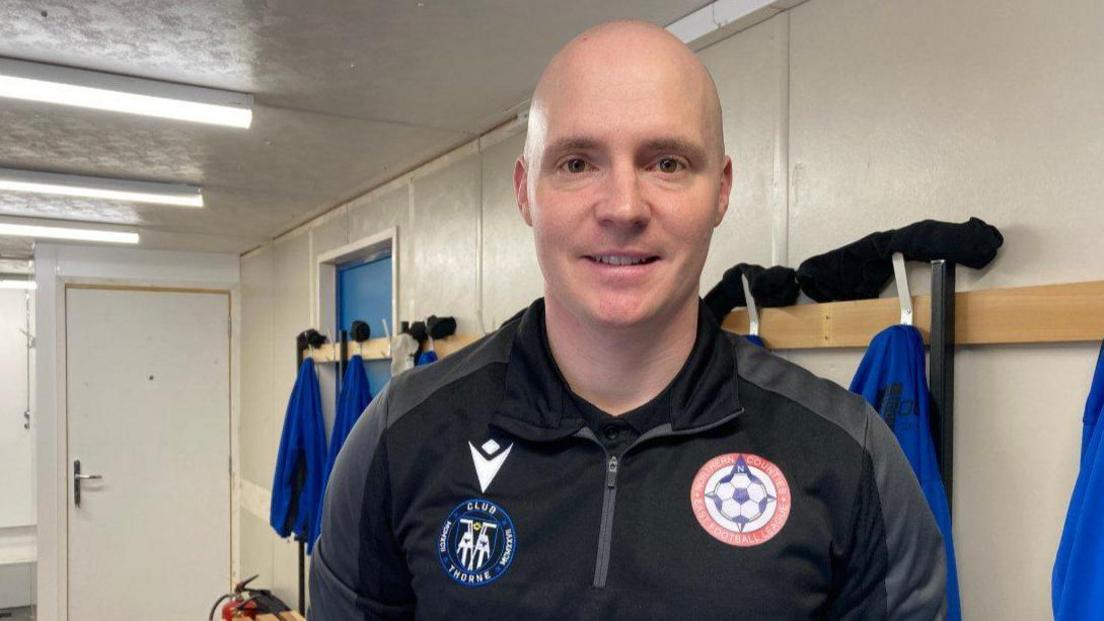 A picture of James O'Neil. He is looking straight ahead and is wearing a club football top and is standing in the new changing rooms. He has a bald head.