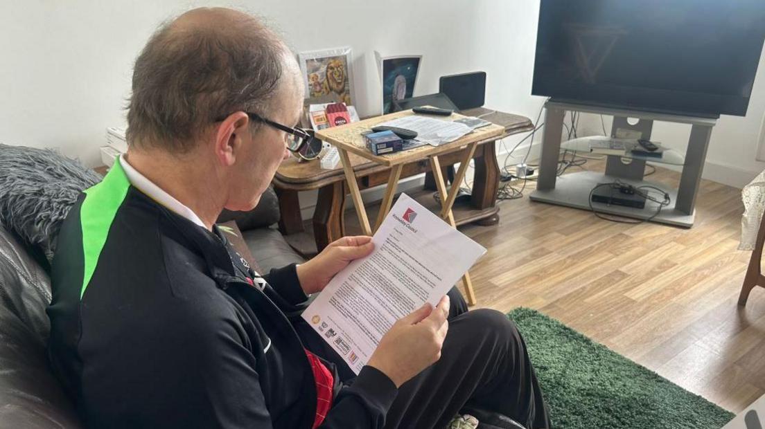 Mr Penfold-Ivany is sat on a sofa in his living room reading a letter from Knowsley Council
