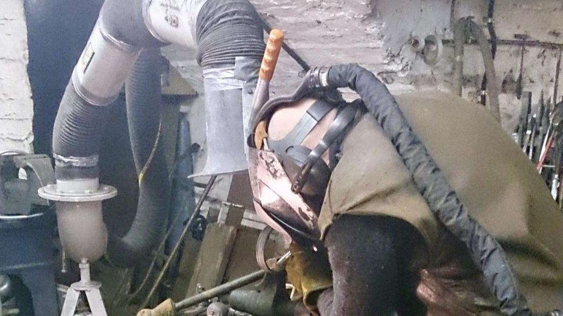 A man hunched over a workbench wearing a visor and industrial protective equipment. Metalworking tools can be seen behind him on the right. On the left there is a long tube and what looks like an extractor flue.