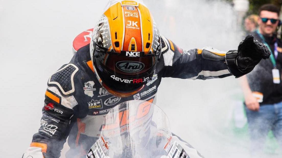 Jamie Williams celebrates with a burnout at the end of the race. He is on his bike with his hand in the air signalling the number one and there is smoke all around him.