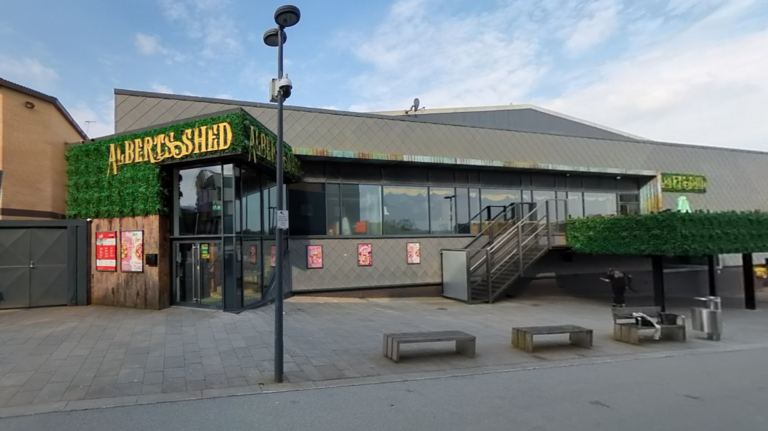 The outside of Albert's Shed Telford. It is a grey building with large glass windows. The sign is on a green leafy background with gold lettering. There is a small staircase up to a deck, with green leaves on its wall