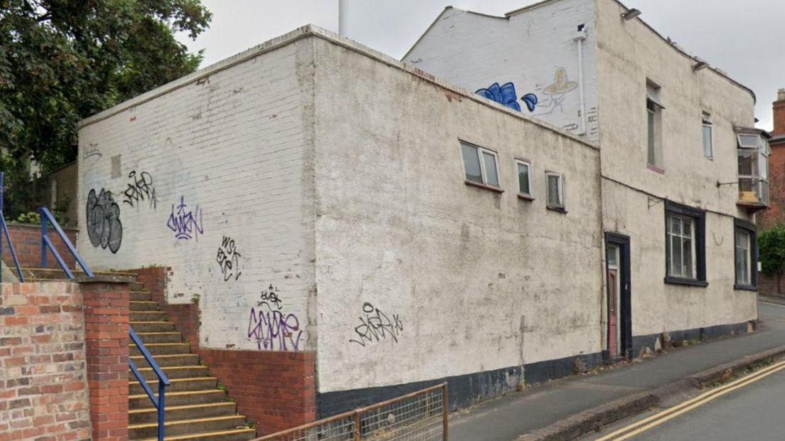 Steps run up the side of the white-painted building which stands on a hill on a street corner in Worcester. There is graffiti on the wall near the steps and on a first-floor wall.