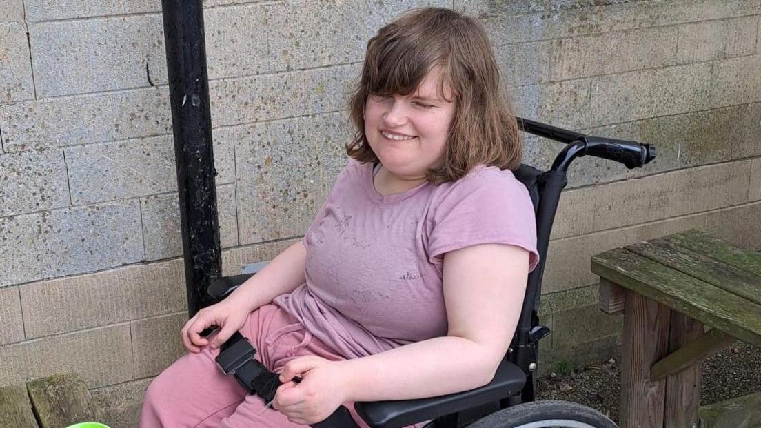 Rhiannon Lucas wearing a lilac T-shirt and a pair of pink tracksuit bottoms. She has shoulder-length brown hair and is smiling at something to the left of the camera. She is sitting in a wheelchair next to a wooden picnic table outside, with a black strap going over her knees.