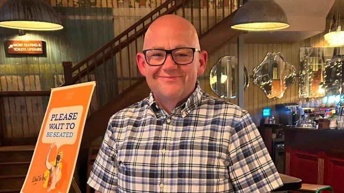 A man in a checked shirt poses and smiles for the camera whilst stood in a bar