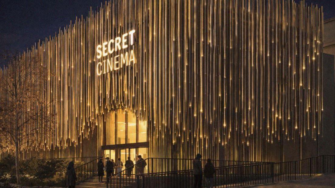 CGI image of the proposed Secret Cinema venue at night. The building is covered with slender vertical metallic rods, illuminated by warm golden lighting. An illuminated "Secret Cinema" sign is positioned above the entrance. People are gathered on the steps outside the venue.