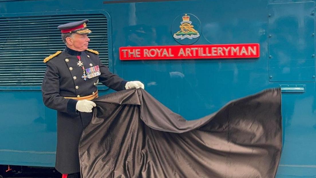 A man in uniform removes a sheet to reveal a plaque with the words "The Royal Artilleryman" affixed to a blue locomotive.