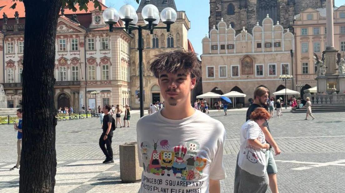 Finn standing in a square in Prague. He is wearing a T-shirt and has his hands behind his back while looking at the camera.