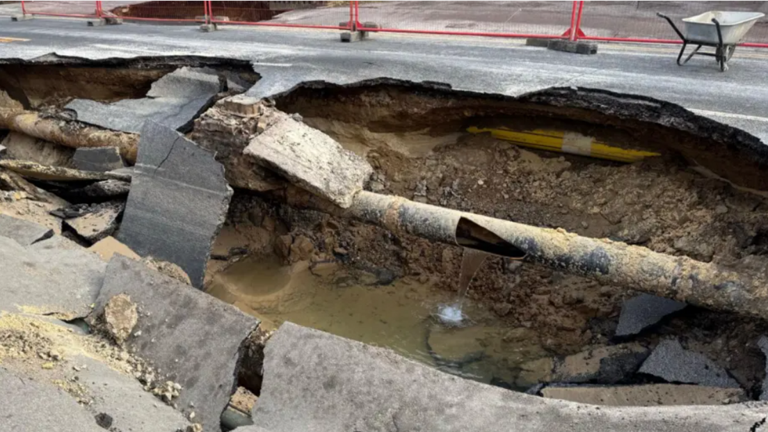 The inside of the sinkhole, as viewed from surface level. The ground beneath the road surface is visible and water flowing out of a hole in a broken pipe is flowing into a pool at the bottom of the sinkhole.