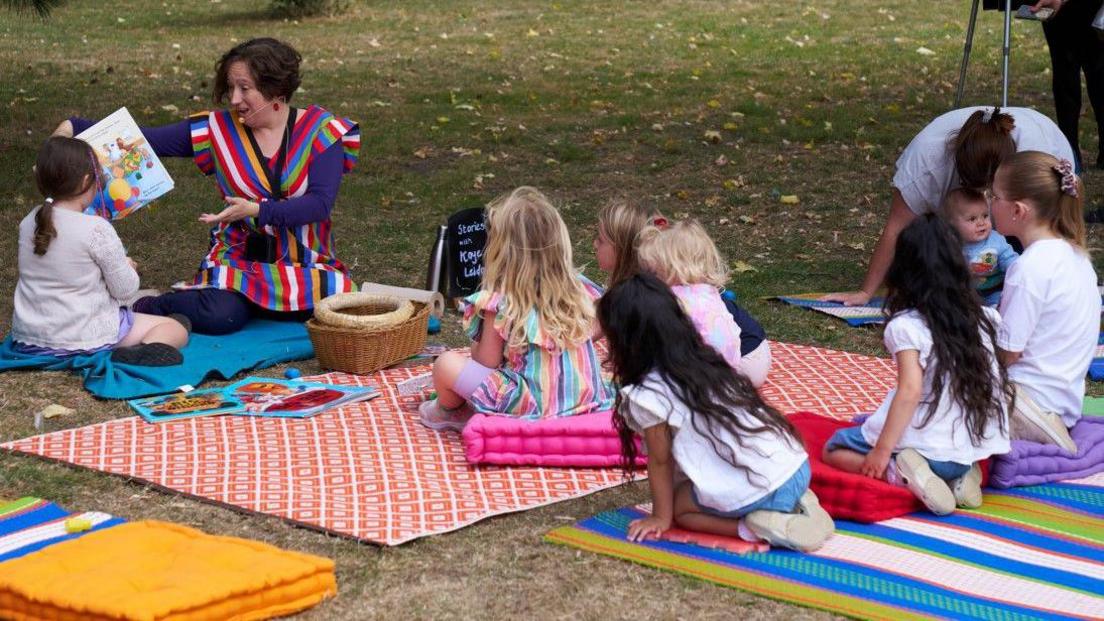 Kaye Mahoney is sitting on a blanket on the grass, with about six children watching on, also sitting on blankets. Kaye is wearing a colourful dress and has a microphone strapped to her cheek.