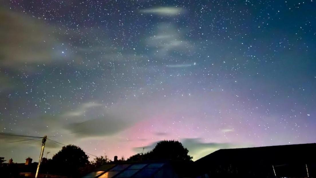 A night-time image of a clear starry sky above rooftops. There is a definite pink hue to much of the sky caused by the northern lights.