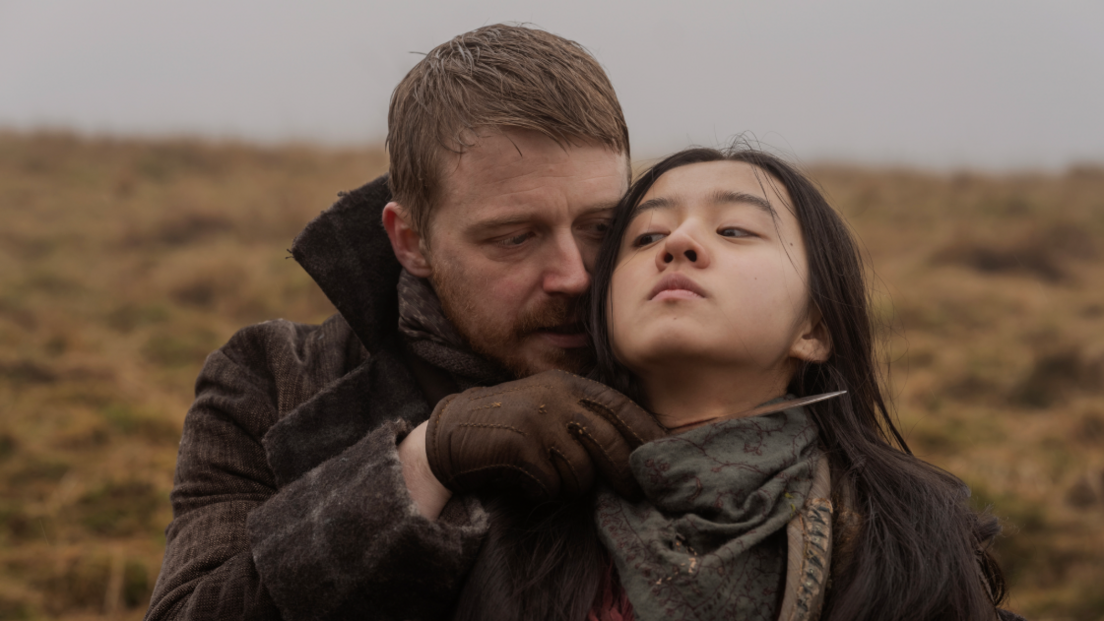 A man with short hair and a beard wearing a brown coat holds a knife to the throat of the lead character in the film Tornado