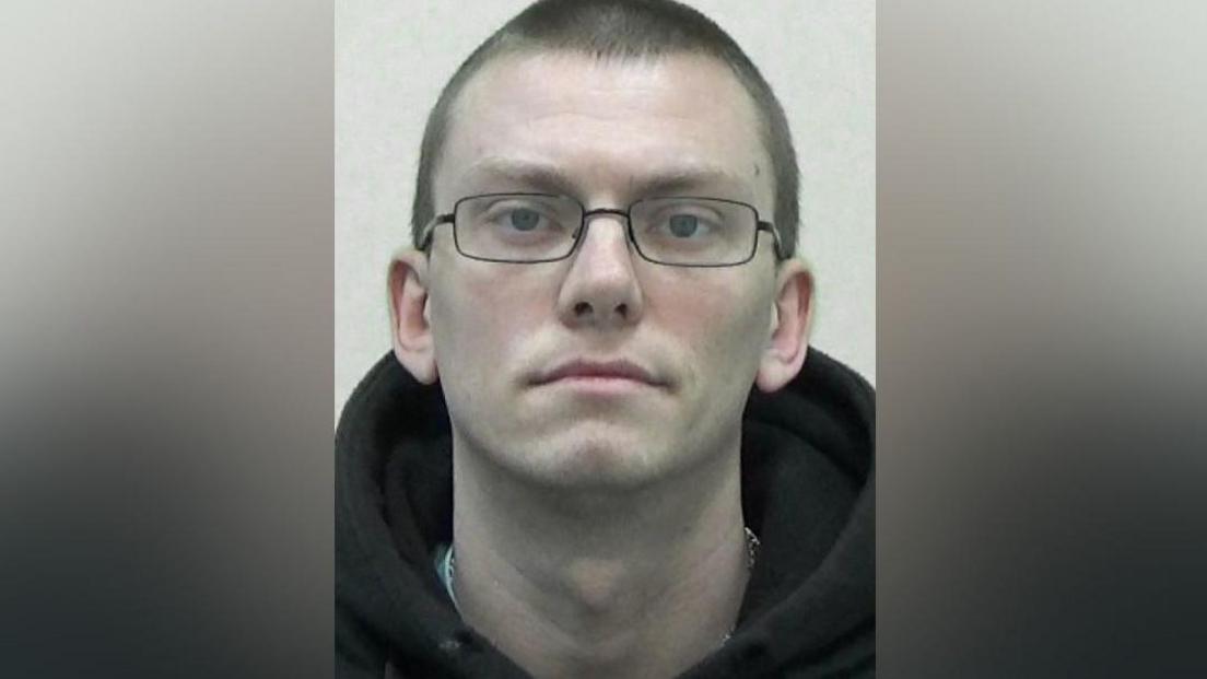 Mugshot of James Gregory. He is clean shaven with short dark hair and is wearing glasses and a black hoody.