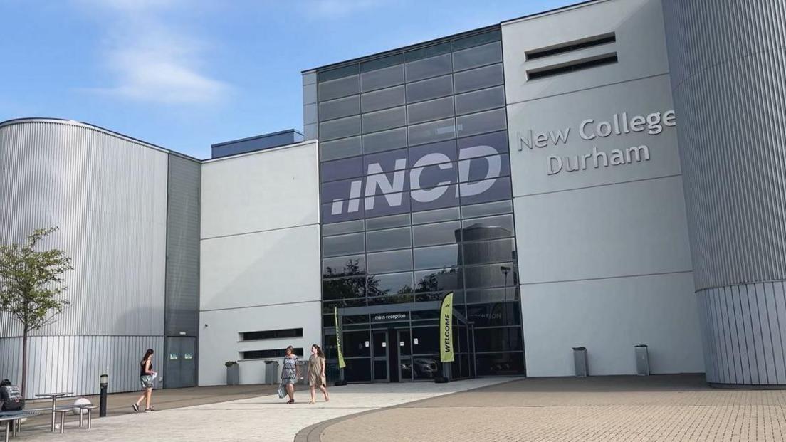 The outside of New College Durham a modern, silver grey rounded building with a column of dark glass tinted windows. There is a white path at the front and three people walking by. 