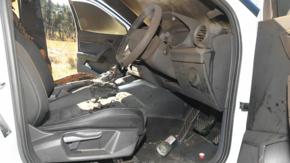 Interior of a white car. It is all covered in soot and the front seats have been burned. An empty glass bottle is in the footwell near the pedals.