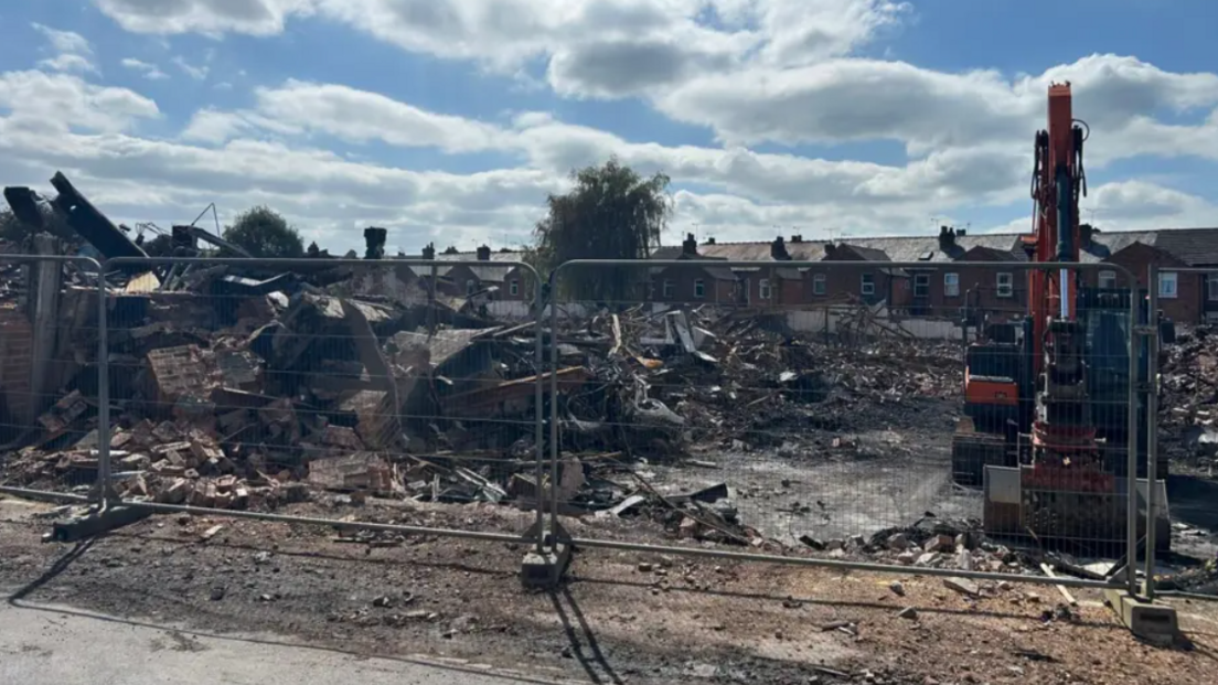 Temporary metal fencing stands in front of piles of rubble and bricks, with a digger seen on the right hand side of the picture. The back of a row of terraced houses run behind the site.