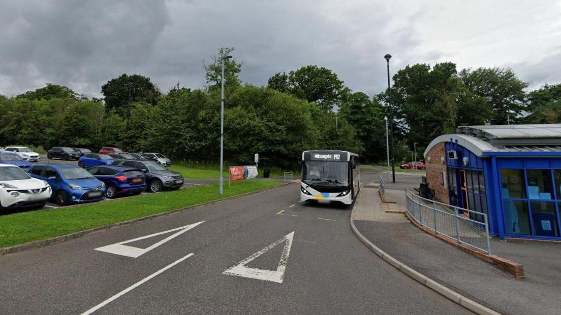 Howlands Park and Ride. There is a blue bus shelter to the right. A white park and ride bus is driving into the car park from the main road. There are parked cars to the left. 