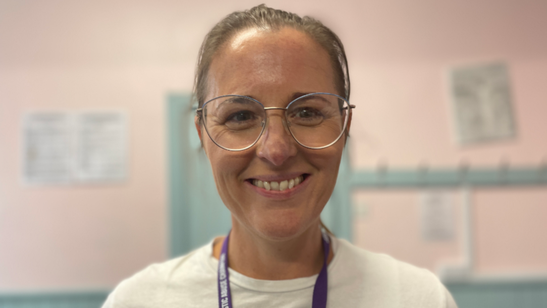 A woman is looking into the camera and smiling. She has light brown hair back in a pony tail and is wearing glasses.