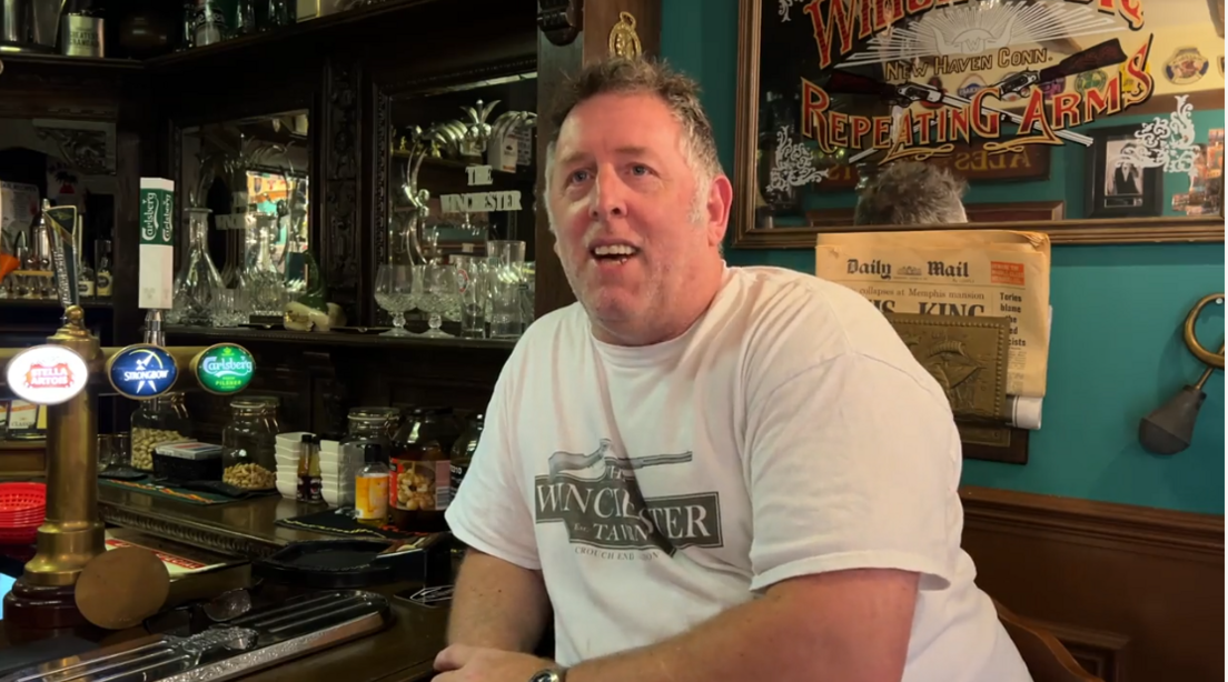 Mr Marchant with grey hair wearing a white t-shirt saying 'The Winchester Tavern' on it, leaning on the bar and smiling at the camera