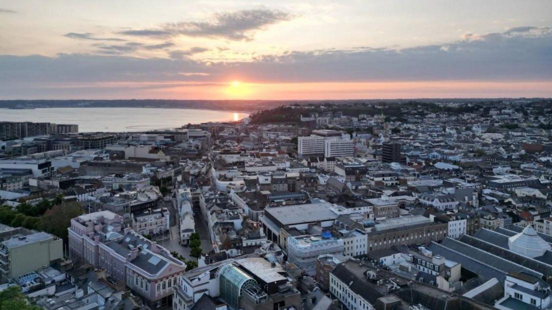 Aerial photo of St Helier, Jersey. Sun appears to be setting or rising over the town. Buildings can be seen beneath the camera with the sea in the top left corner.