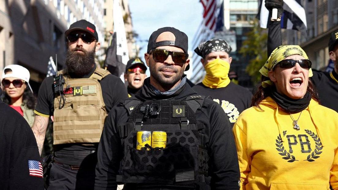 Tarrio in sunglasses and a vest containing two yellow cans, flanked by others wearing Proud Boys gear