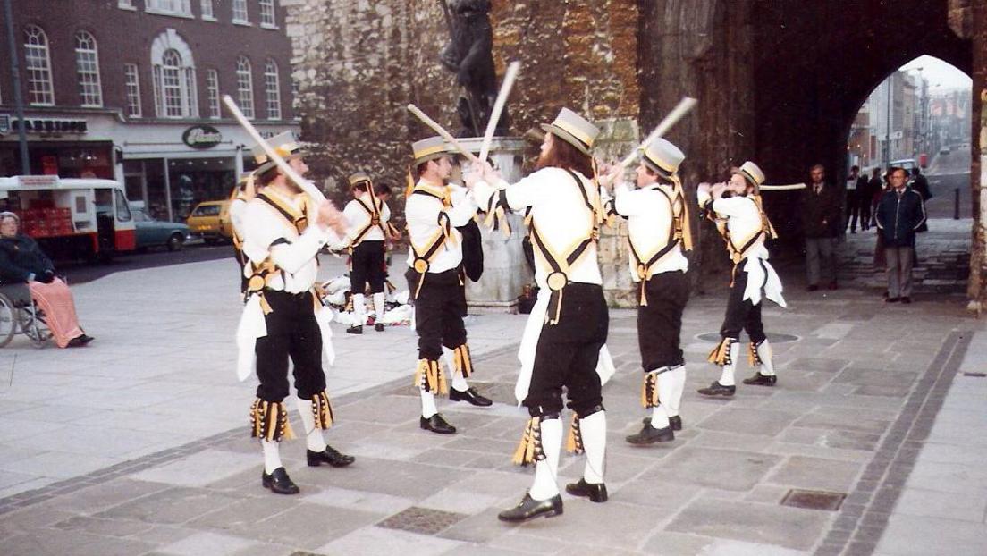 Five Morris men from King John's Morris Men wearing top hats white shirts, black trousers and white socks pulled up to the knee. They have yellow and black tassels and accessories. They are dancing in front of the bargate in Southampton at dawn