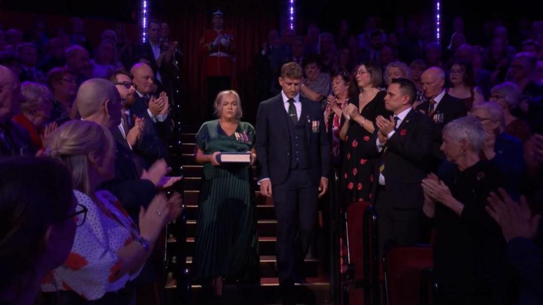 Hayley Lawrence is carrying a book and is wearing a green dress and military medals, Alex Lowe wears a suit and medals, they are walking down stairs, while the audience claps 