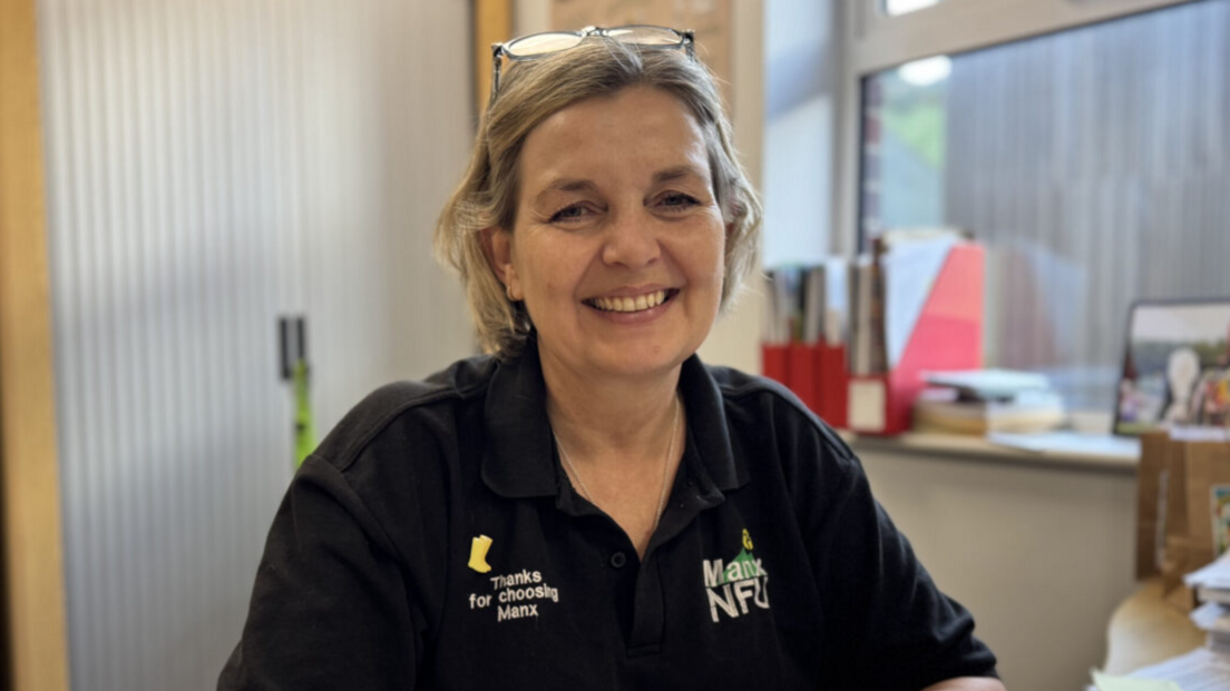 Sarah Comish has short blonde hair and is wearing a black polo shirt with the Manx NFU logo on the front. She's smiling, sitting in her office with a window to the right and a storage unit behind her on the left.