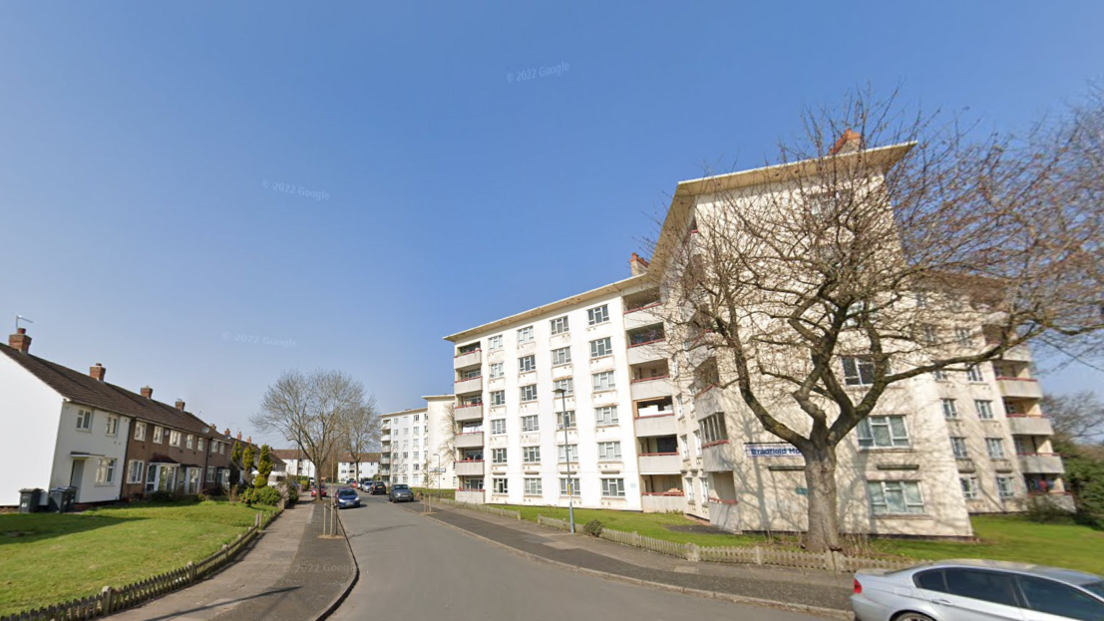 A six-storey block of flats, painted white, opposite terraced houses in a small road on a sunny day.