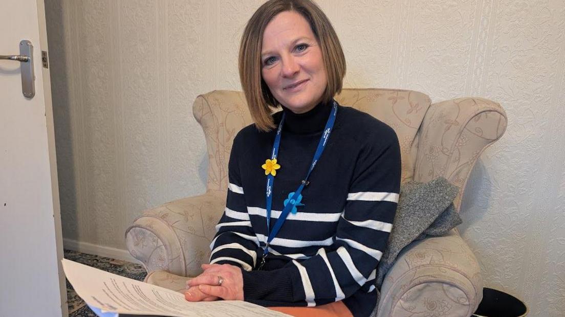 A smiling woman sat in an armchair holding a piece of paper. She has shoulder length brown hair and is wearing a navy blue and white striped jumper. She has a lanyard with a yellow daffodil attachment around her neck. 