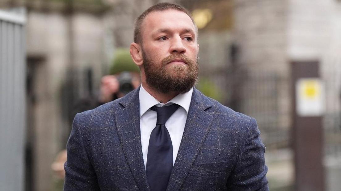 Conor McGregor walks outside court. He has cropped, light brown hair and and a beard. He is wearing a checked blue suit, light shirt and navy tie and is looking to his side. 