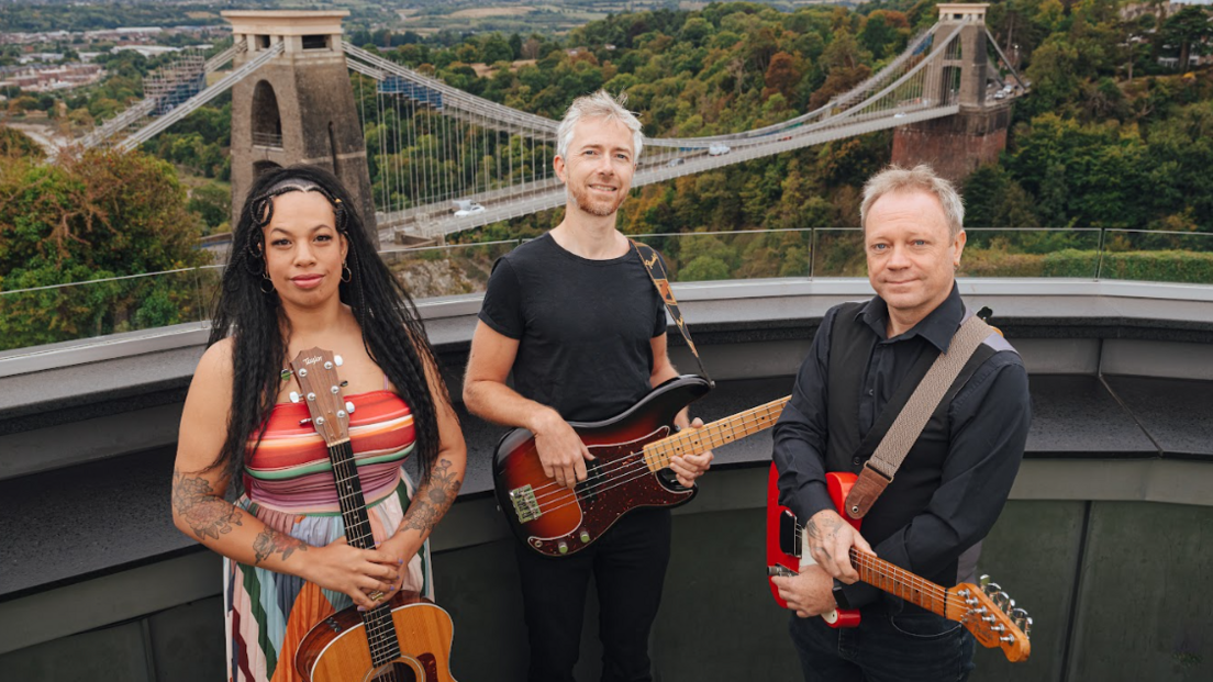 Three members of a band standing on a balcony overlooking the Clifton Suspension Bridge. Lady Nade is holding a classical guitar - she has a multi-coloured dress on and is smiling to camera. The other band members are male, both also holding electric guitars