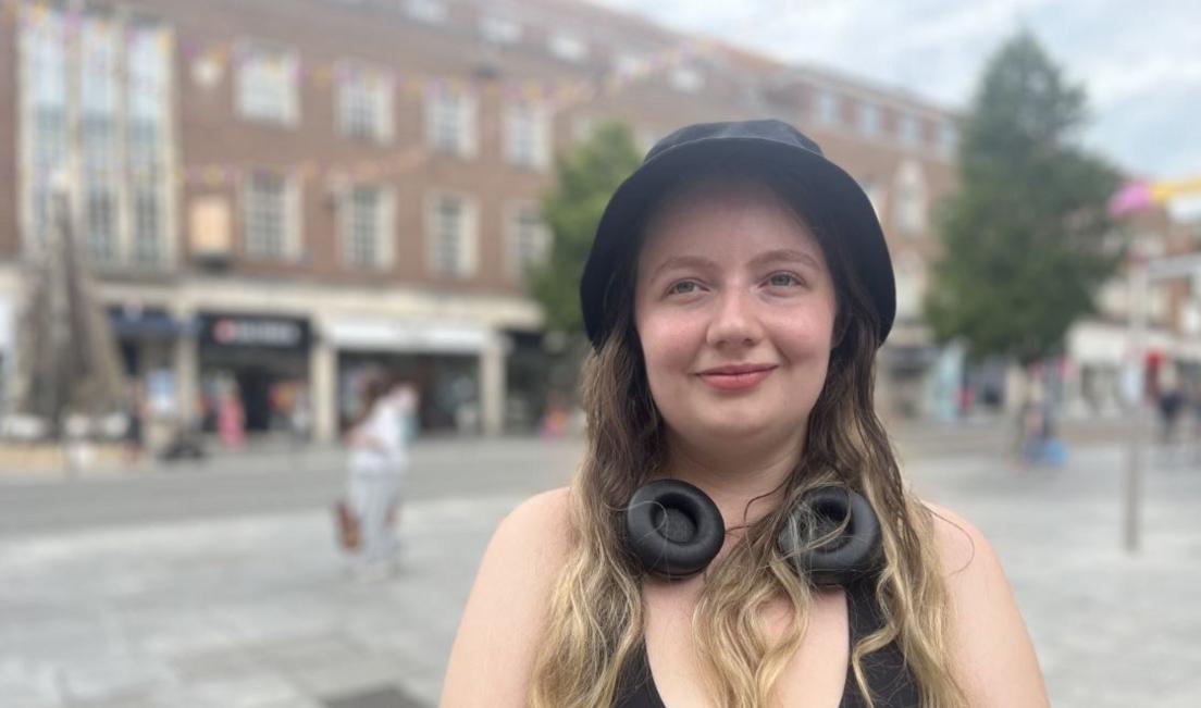 Kat Smith, standing in a street in Exeter city centre. She has a black hat and has long hair with blonde highlights. She is wearing a black top and also has a pair of black headphones around her neck.