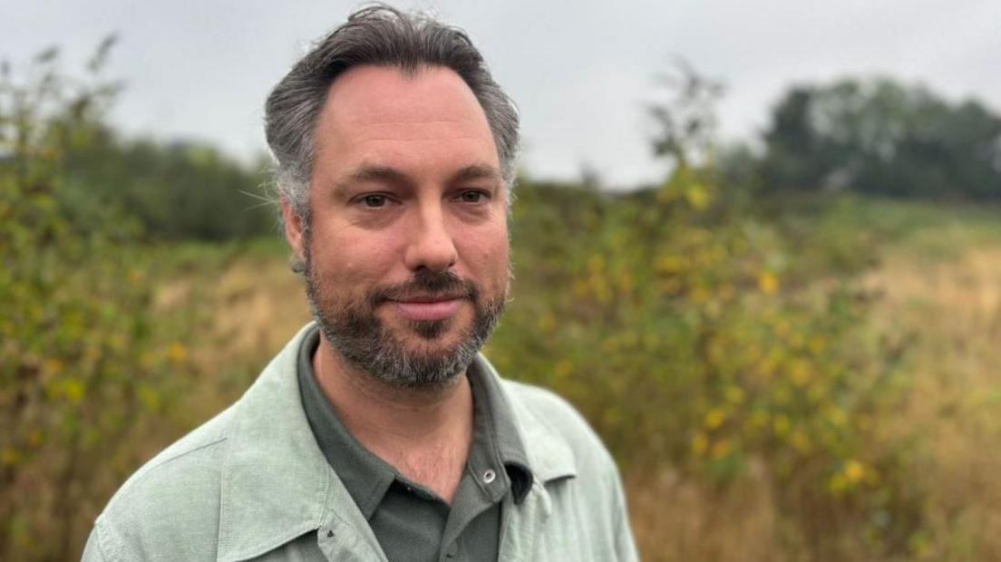 A man with dark greying hair and beard wearing a khaki shirt and green linen jacket. He is standing in a field, with yellow flowers on shrubs in the background.