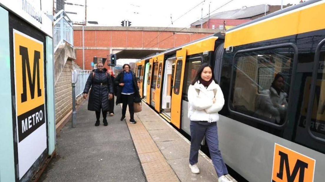 Passengers walk along a Metro platform at Millfield station next to the new train. It has a yellow-and-grey livery with the Metro logo on the side of one of the carriages.