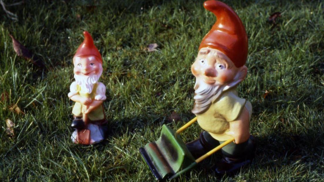 Two garden gnomes, with pointed red hats and sporting long white beards, are pictured "holding" tools including a spade and a lawnmower.