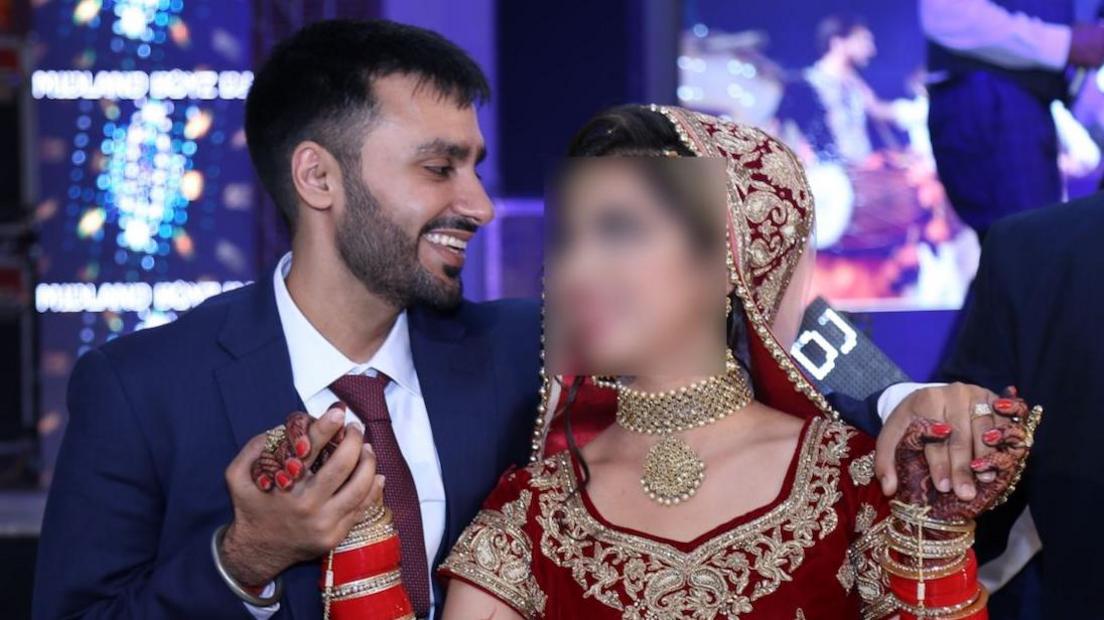 Jagtar Singh Johal wears a navy suit with a dark red tie and white shirt and holds Gurpreet Kaur's hands as they dance. She wears a red sari with gold embroidery and a gold necklace.