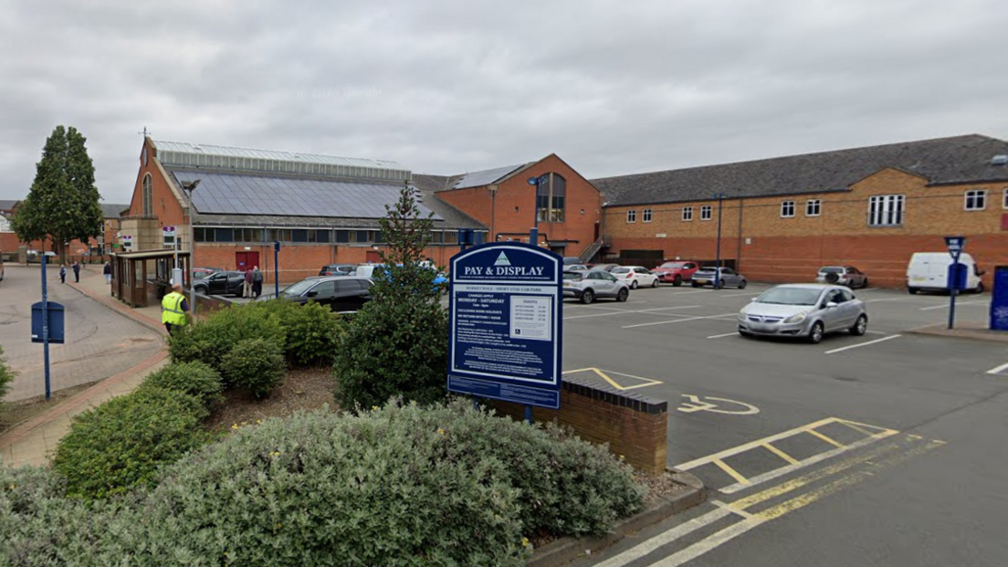 A council car park in Market Harborough next to the town's market hall. A number of cars are parked in spaces, and there is a shrubbery bed just outside the car park, and a large Pay & Display sign can be seen.