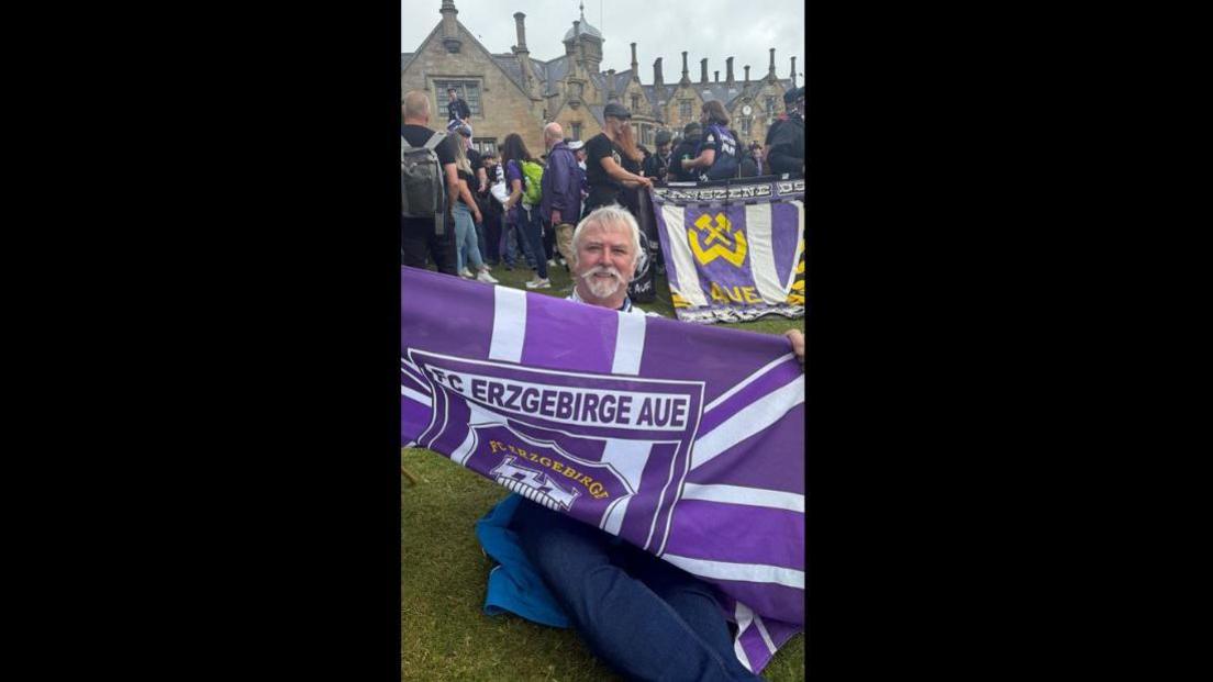 A football fan holds the flag of the team Erzgebirge Aue. He is sitting on the grass, with fellow fans in the background. He has grey hair and a beard. 