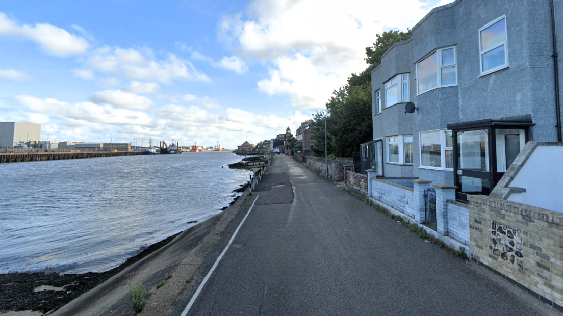 A google street view image of Riverside Road in Gorleston. The River Yare is one side of the road and riverside homes painted grey are on the other.
