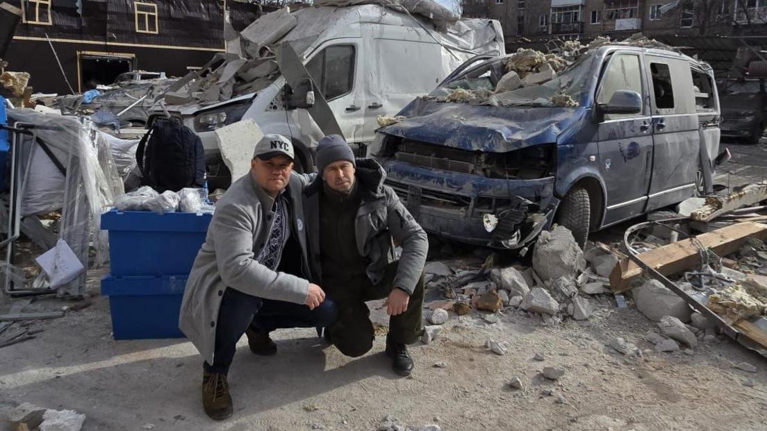 Karol Swiacki is crouching next to Marc Edwards, surrounded by the rubble and impact of the missile strike. Karol wears a grey NYC cap with a grey woolen coat, patterned t shirt and jeans with brown boots. Marc wears a slate grey beanie, with a grey puffer coat, green combat trousers and black trousers.